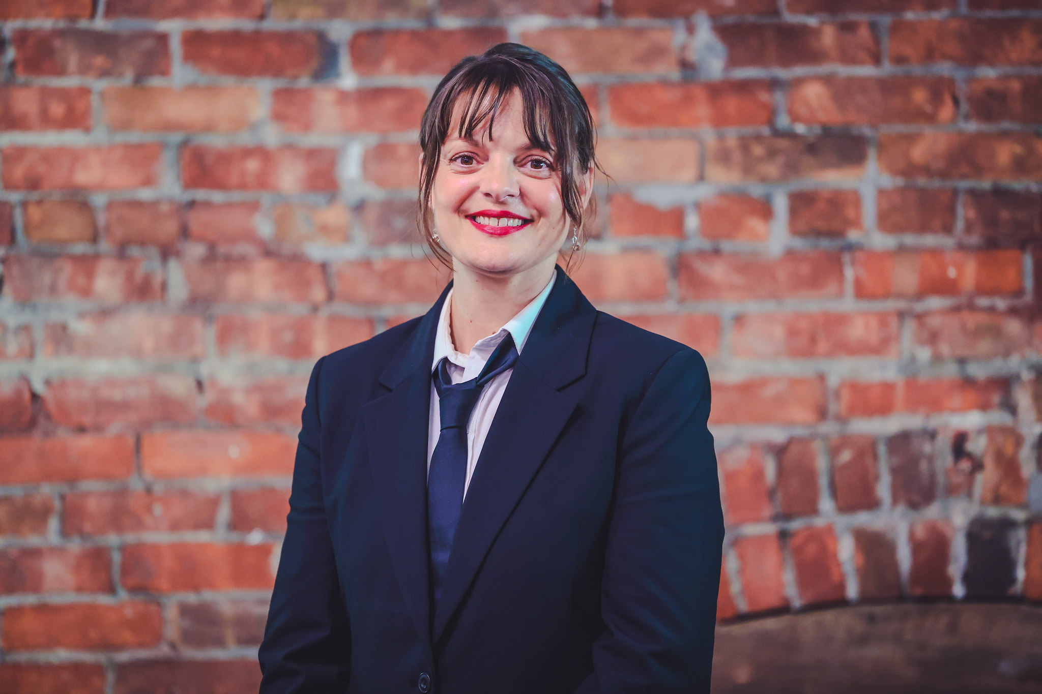 A young woman wearing a suit and tie smiles in front of a brick wall