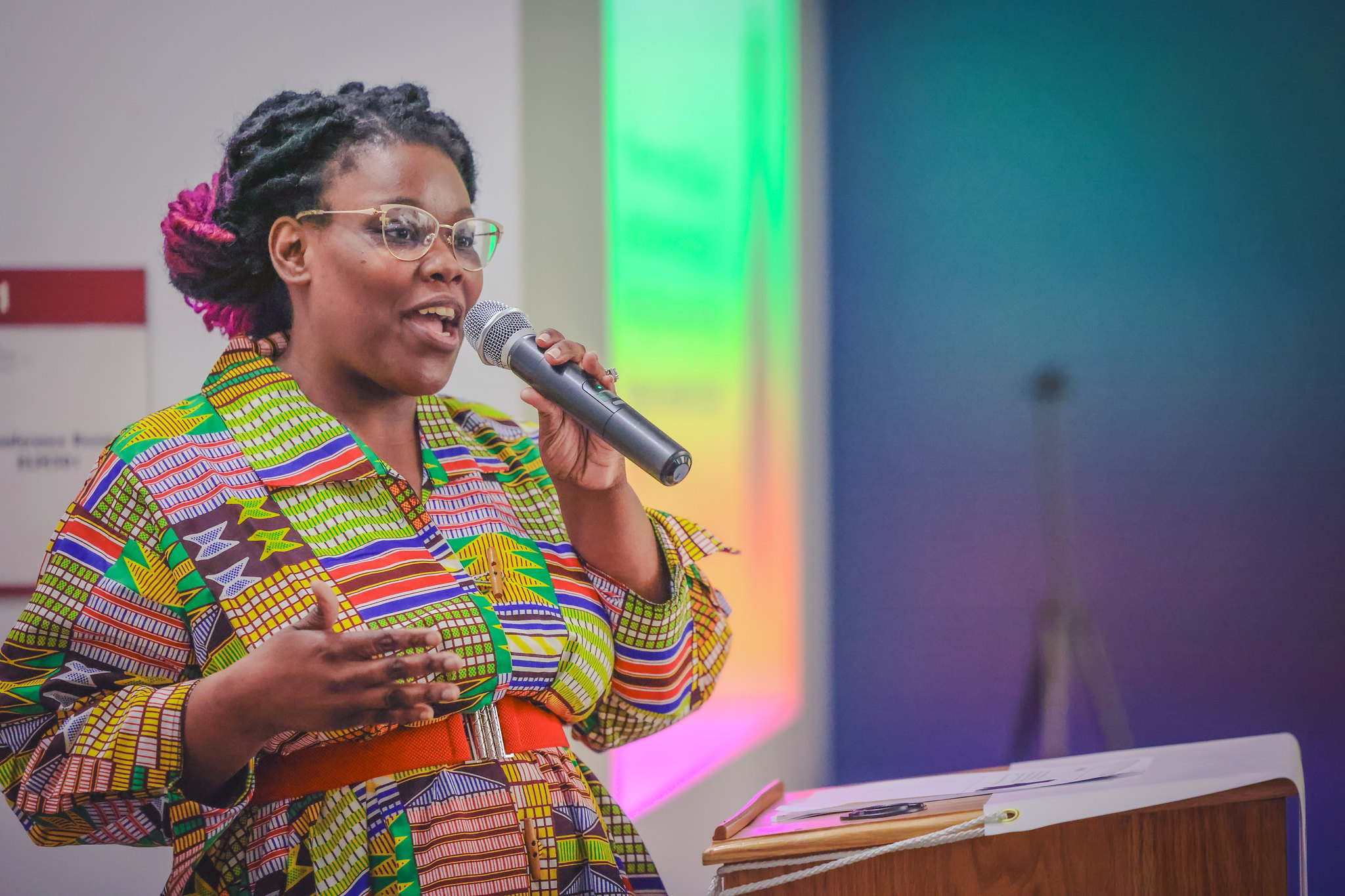 A Black woman with glasses, braided hair, and a colorful dress speaks into a microphone at the 2024 CoStarters graduation
